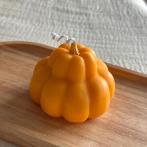 May include: A pumpkin-shaped candle in a vibrant orange color with a smooth, segmented surface. The candle features a small, metallic gold-colored stem at the top center, from which a white, coiled cotton wick extends upwards.