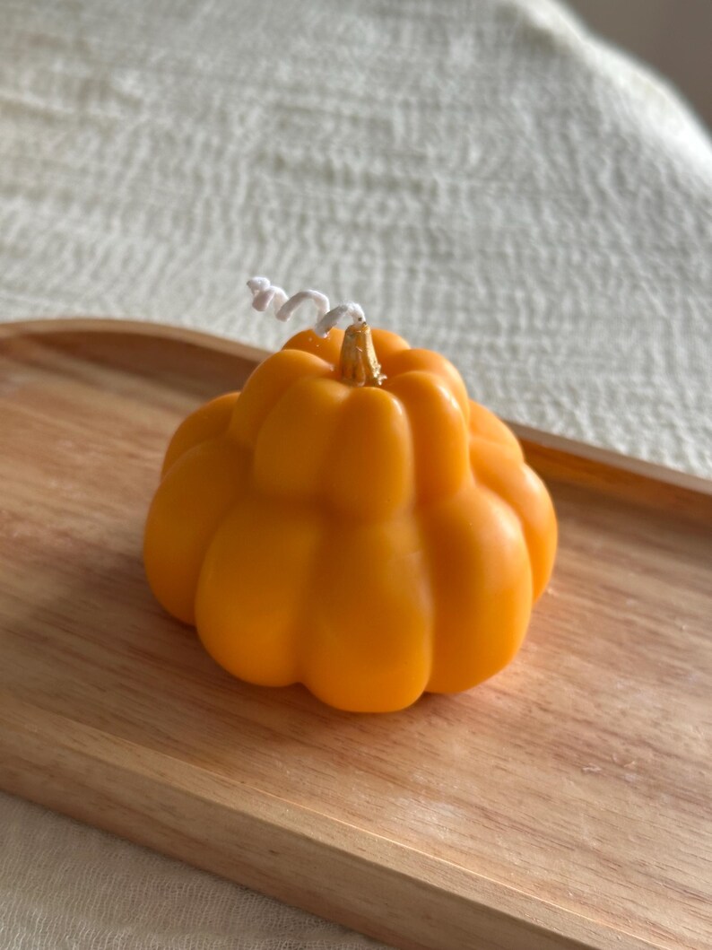 May include: A pumpkin-shaped candle in a vibrant orange color with a smooth, segmented surface. The candle features a small, metallic gold-colored stem at the top center, from which a white, coiled cotton wick extends upwards.