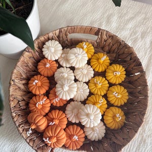 May include: A collection of small, pumpkin-shaped candles in shades of white, orange, and yellow, each featuring a central white wick. These wax candles are arranged together inside a round, woven brown wicker basket with two side handles.