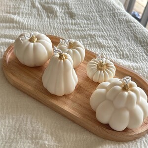May include: Five off-white pumpkin-shaped candles with gold-painted stems and white wicks, arranged on an oval wooden tray. The candles vary in size and shape, featuring ribbed textures that mimic the appearance of small gourds.