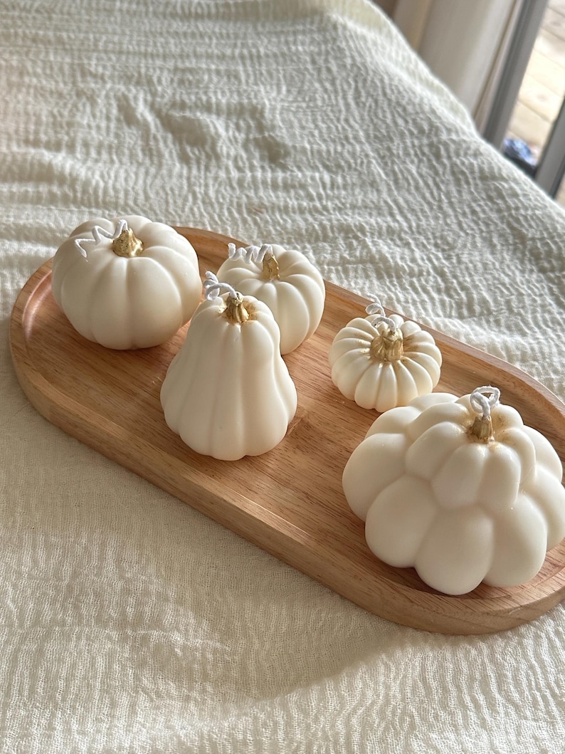 May include: Five off-white pumpkin-shaped candles with gold-painted stems and white wicks, arranged on an oval wooden tray. The candles vary in size and shape, featuring ribbed textures that mimic the appearance of small gourds.