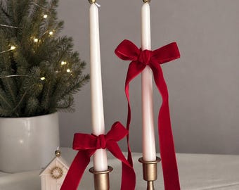 Puede incluir: Dos velas cónicas blancas colocadas en candelabros de metal dorado cepillado. Cada vela está adornada con una cinta larga de terciopelo rojo intenso atada en forma de lazo. Las velas tienen una pequeña cuenta dorada en la punta de la mecha.