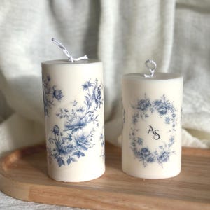 May include: Two ivory pillar candles with blue floral patterns. One candle has a full floral design, the other a floral wreath with the initials "AS". Both have white wicks and are on a wooden tray. The candles are approximately 7 cm in diameter.