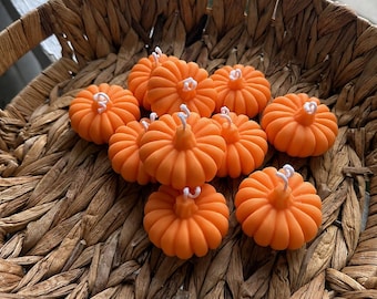 Puede incluir: Una colección de pequeñas velas de color naranja brillante con forma de calabaza y mechas blancas, dispuestas en una cesta redonda de mimbre marrón. Cada vela presenta una superficie redondeada y acanalada con un tallo central para la mecha.