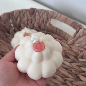 May include: A cream-colored, cloud-shaped candle with a rounded, multi-lobed surface. A small, pink, pumpkin-shaped metal charm with gold-colored star accents is attached to the white wick at the top center of the candle.