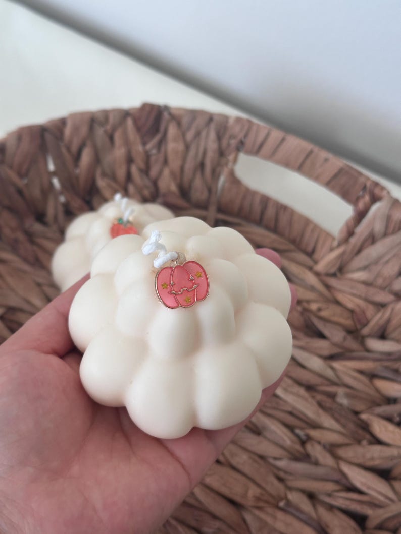 May include: A cream-colored, cloud-shaped candle with a rounded, multi-lobed surface. A small, pink, pumpkin-shaped metal charm with gold-colored star accents is attached to the white wick at the top center of the candle.