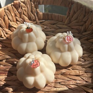 May include: Three off-white, pumpkin-shaped candles with a rounded, segmented design. Each candle features a single white wick topped with a small, colorful metal pumpkin charm. The charms are orange, pink, and light pink with star details.