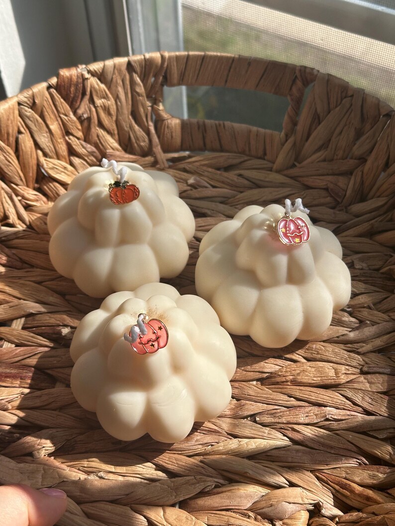 May include: Three off-white, pumpkin-shaped candles with a rounded, segmented design. Each candle features a single white wick topped with a small, colorful metal pumpkin charm. The charms are orange, pink, and light pink with star details.