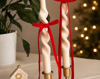 Puede incluir: Dos velas cónicas en espiral de color blanco roto en candelabros de metal dorado, cada una adornada con un lazo de terciopelo rojo y una pequeña campana dorada en la parte superior. A la izquierda hay una vela pequeña en forma de casa con detalles dorados.