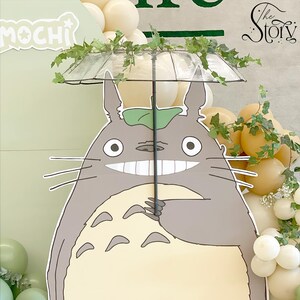 May include: A large, smiling cartoon character with a clear umbrella decorated with greenery. The character is gray and cream-colored, with large eyes and whiskers. Balloons and decorative foliage are in the background.