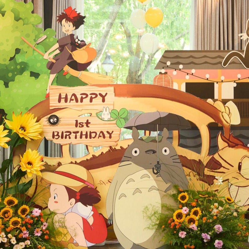 May include: A colorful birthday party scene featuring characters from the animated film "My Neighbor Totoro." The scene includes a sign that says "Happy 1st Birthday," sunflowers, and other decorative elements. The characters include Totoro and Kiki.