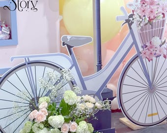 Puede incluir: Un recorte decorativo de bicicleta con una cesta llena de flores rosas. La bicicleta es azul claro y blanca, con un asiento y manillar a juego. El fondo incluye globos y el texto "The Story".