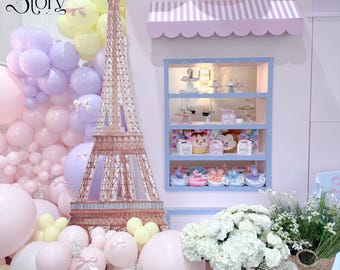 Puede incluir: Una exhibición decorativa con una combinación de colores pastel, con globos rosas, amarillos y morados. Una torre Eiffel de cartón se encuentra frente a una vitrina con postres. El texto "The Story" está en la esquina superior izquierda.