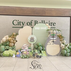 May include: A whimsical baby shower display featuring a large Totoro cutout, balloons in shades of green and beige, and character cutouts. The backdrop includes the text "City of Bellaire" and "Baby Mochi".