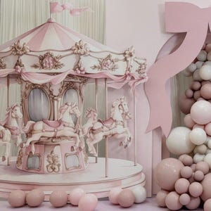 Puede incluir: Un carrusel rosa y blanco con tallas detalladas y caballos. El carrusel tiene un dosel a rayas y está decorado con cintas y detalles florales. Un gran lazo rosa y un grupo de globos también están presentes.
