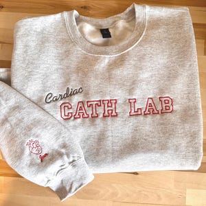 May include: A light gray sweatshirt with the words "Cardiac CATH LAB" embroidered in red and black. The left sleeve features a red embroidered heart and the name "Jon". The sweatshirt is made of a soft material.