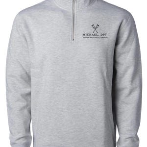 May include: A light gray sweatshirt with a quarter-zip neckline. The sweatshirt features the text "MICHAEL, DPT DOCTOR OF PHYSICAL THERAPY" in black, along with a graphic of crossed crutches. The long-sleeved top has ribbed cuffs and a ribbed hem.