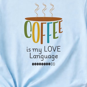 Puede incluir: Una camiseta de manga corta de color azul claro con un gráfico de una taza de café marrón con vapor saliendo. El texto dice COFFEE is my LOVE Language, seguido de una fila de pequeños iconos de granos de café negros al frente.