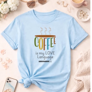 Könnte beinhalten: Hellblaues T-Shirt mit hochgekrempelten Ärmeln. Der Aufdruck "COFFEE is my LOVE Language" in bunten Buchstaben, mit einer Kaffeetasse darüber. Ein lässiges Kleidungsstück.