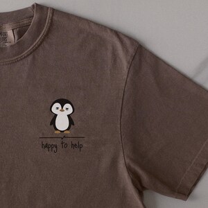 Könnte beinhalten: Ein braunes T-Shirt mit kurzen Ärmeln und einer kleinen Grafik eines Pinguins mit müdem Gesichtsausdruck. Unter dem Pinguin befinden sich eine horizontale Linie und der Text "happy to help" in einer einfachen, dunklen Handschrift-Schriftart.
