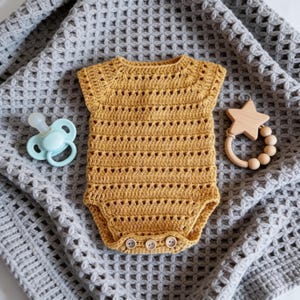 May include: A mustard yellow crochet baby bodysuit with short sleeves and three small buttons at the bottom. To the left is a light blue dummy, and to the right is a wooden star-shaped rattle with round beads, all resting on a grey waffle-knit blanket.