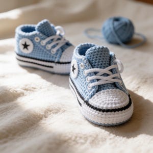 May include: A pair of light blue crochet baby trainers featuring white laces, white toe caps, and a black stripe along the sole. Each shoe displays a circular white patch on the side with a black star emblem, accompanied by a matching ball of yarn.