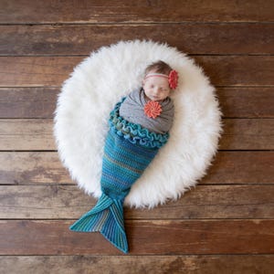 May include: A crochet mermaid tail cocoon in shades of teal, blue, and green with a ruffled waist. The set includes a matching headband and a decorative pin, both featuring a coral-colored flower, worn by a swaddled infant on a white rug.