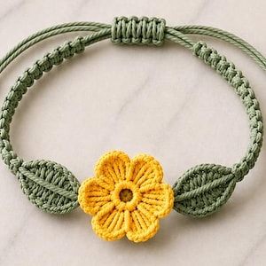 May include: A macrame bracelet featuring a central yellow flower motif flanked by two green leaf shapes. The band is constructed from sage green cord with a sliding knot closure, creating an adjustable fit. The design is symmetrical and detailed.