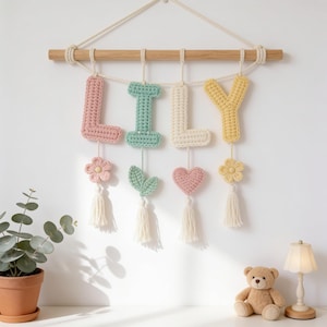 May include: A wall hanging featuring the name LILY spelled out in crocheted letters suspended from a wooden rod. The letters are pink, mint green, cream, and yellow, each accented with a small crocheted flower, leaf, or heart and a white tassel.