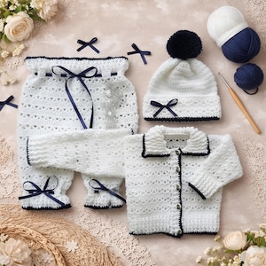 May include: A white crochet baby outfit set with navy blue trim and bows. The set includes pants, a long-sleeved jacket, and a beanie with a navy pom-pom. The outfit is displayed with yarn, a crochet hook, and decorative bows.