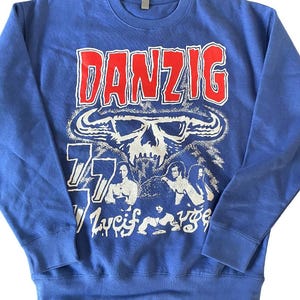 May include: Blue sweatshirt with the word "DANZIG" in red block letters above a white skull graphic. Below the skull are the numbers "77" and the words "Lucifuge". The sweatshirt has a crew neck and long sleeves.