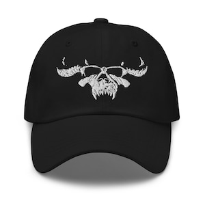 May include: Black baseball cap with a white embroidered skull design. The skull features horns, a nose, and sharp teeth. The cap has a curved brim and a button on top.