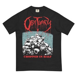 May include: Black t-shirt featuring the band name "Obituary" in red, dripping font above a pile of skulls. Below the skulls, the text "Chopped in Half" is printed in white. The shirt has a classic crew neck.