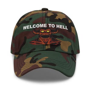 May include: Camouflage baseball cap with the text "WELCOME TO HELL" in white. The cap features a red and orange illustration of a horned demon with yellow eyes.