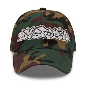 May include: A camouflage baseball cap with the white embroidered word "Dystopia" in a graffiti-style font. The cap features a curved brim and a black button on top. The fabric has a pattern of green, brown, and black.
