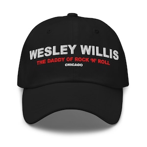May include: Black baseball cap with the embroidered text "WESLEY WILLIS" in white, "THE DADDY OF ROCK 'N' ROLL" in red, and "CHICAGO" in white. The cap has a curved brim and a button on top.