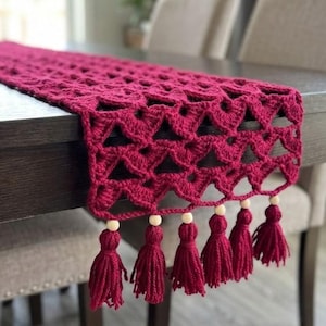 Può includere: Runner da tavolo all'uncinetto bordeaux con motivo geometrico e nappe. Il runner è drappeggiato su un tavolo in legno scuro, mettendo in mostra il suo design intricato e la frangia decorativa. Le nappe sono rifinite con perline di legno.