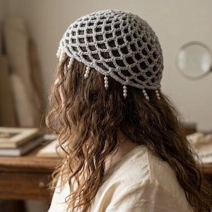 Puede incluir: Un gorro de crochet gris con un diseño de red, adornado con cuentas de perlas colgantes. El gorro lo lleva una persona con cabello castaño largo y ondulado. La imagen está tomada en interiores, con una mesa de madera y otros objetos en el fondo.