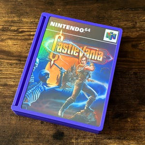 Peut inclure: Un boîtier de jeu Nintendo 64 violet avec la jaquette du jeu Castlevania. L'illustration représente un guerrier avec une épée, un squelette et un château. Le titre du jeu est en or.
