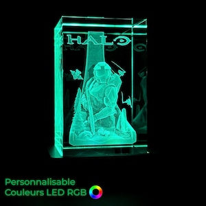 Può includere: Una lampada LED rettangolare in acrilico che emette una luce verde brillante. Il pannello frontale presenta un design inciso di un soldato in armatura con la parola HALO in alto. La scena include un paesaggio con alberi e un piccolo veicolo.