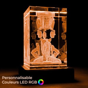 Op de afbeelding: Een rechthoekige, verlichte glazen sculptuur met een oranje gloed, met een gedetailleerde 3D-gravering van een figuur en machines. De sculptuur staat op een zwarte basis. De tekst "Personnalisable Couleurs LED RGB" is zichtbaar.