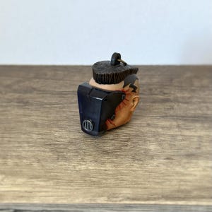 Puede incluir: Una figura en miniatura de una cabeza con una máscara azul oscuro y un peinado marrón. La cabeza es de color canela con marcas rojas. La máscara tiene un diseño circular en el lateral. La figura está sobre una superficie de madera.