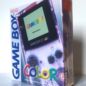 Puede incluir: Una caja de consola de videojuegos Game Boy Color. La caja presenta un diseño púrpura transparente de la consola con las palabras "GAME BOY COLOR" en letras coloridas. El logotipo de Nintendo es visible.