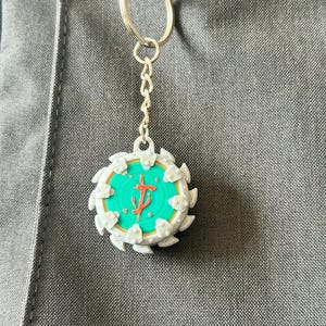 Peut inclure: Un porte-clés avec un pendentif circulaire présentant un motif turquoise et doré avec un symbole rouge. Le pendentif est entouré de formes blanches en forme de dents. Le porte-clés est attaché à une chaîne et un anneau argentés.