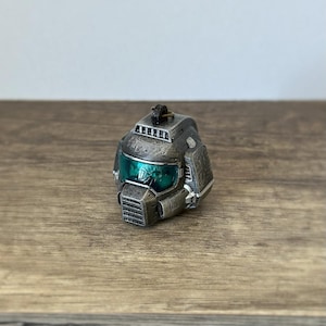 Może przedstawiać: Metalowy szary hełm z zielonym wizjerem, prawdopodobnie miniaturowa replika. Hełm ma szczegółowe panele i małą pętlę na górze. Obiekt jest ustawiony na drewnianej powierzchni.