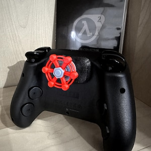 May include: Black video game controller with a red and blue accessory attached. A black collector's edition case with the Half-Life 2 logo is in the background.