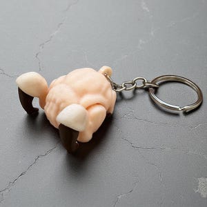 Peut inclure: Un porte-clés en forme de mouton rose clair et texturé, avec des sabots marron foncé et un porte-clés argenté. Le mouton est couché, les pattes repliées sous son corps. Le porte-clés est sur une surface grise.