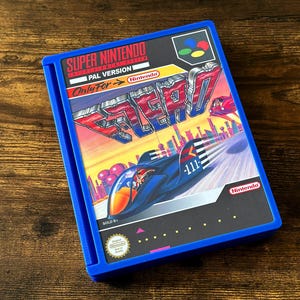 Peut inclure: Un boîtier de cartouche de jeu Super Nintendo bleu avec le jeu F-Zero. La couverture montre une voiture de course futuriste traversant une ville. Le titre du jeu est en rouge et bleu, avec le logo Nintendo.