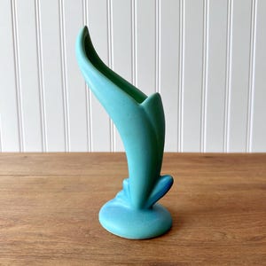 May include: A turquoise ceramic vase with a curved, organic shape. The vase has a flared opening and a rounded base. The color is a solid, matte turquoise. The vase is displayed on a wooden surface.
