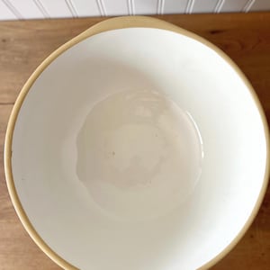 K&ouml;nnte beinhalten: Eine wei&szlig;e Keramiksch&uuml;ssel mit beigem Rand. Die Sch&uuml;ssel hat eine abgerundete quadratische Form und steht auf einer Holzoberfl&auml;che. Das Innere der Sch&uuml;ssel ist cremewei&szlig;.